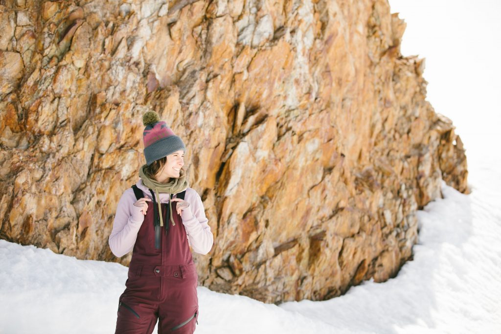 A female skier standing in the snow wearing maroon colored ski bibs