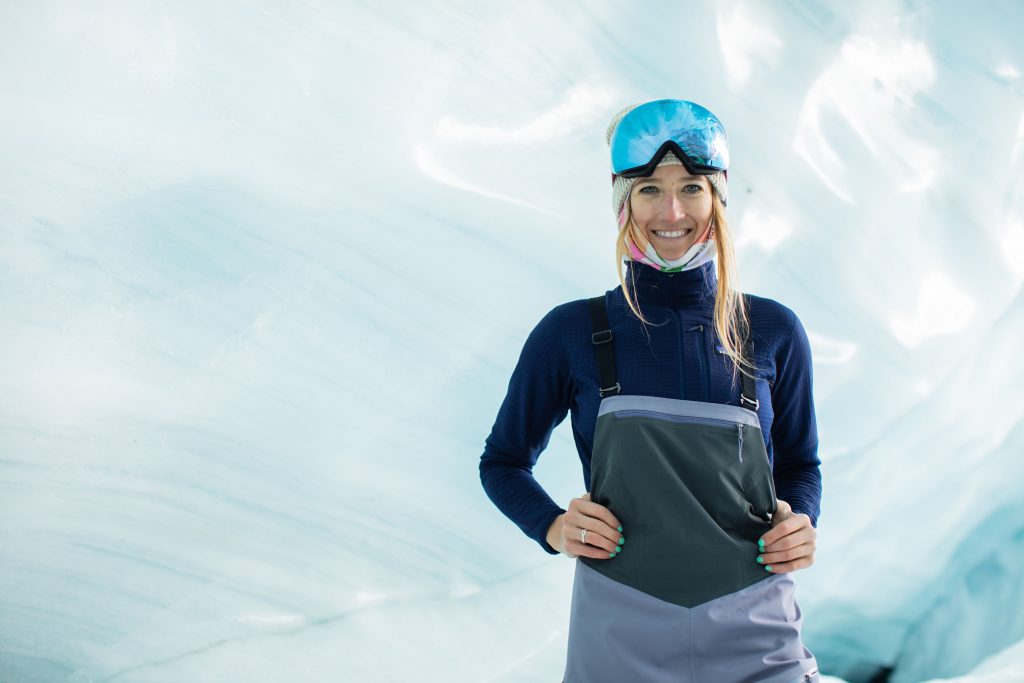 A female skier in front of a glacier wearing ski bibs and goggles