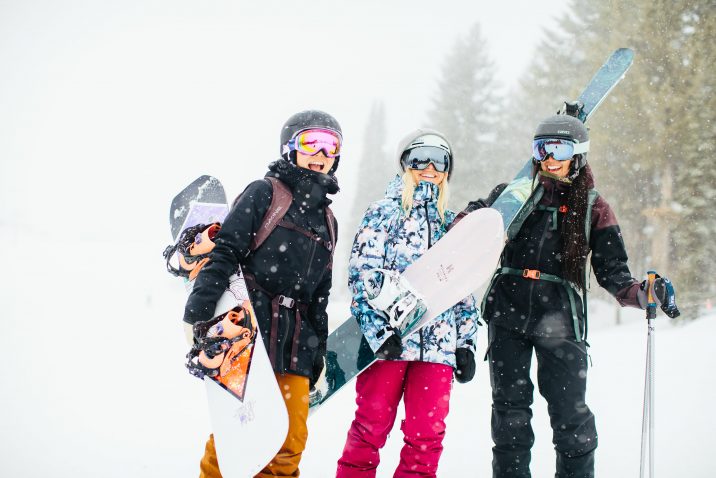 2 female snowboarders and 1 female skier standing together smiling in the snow
