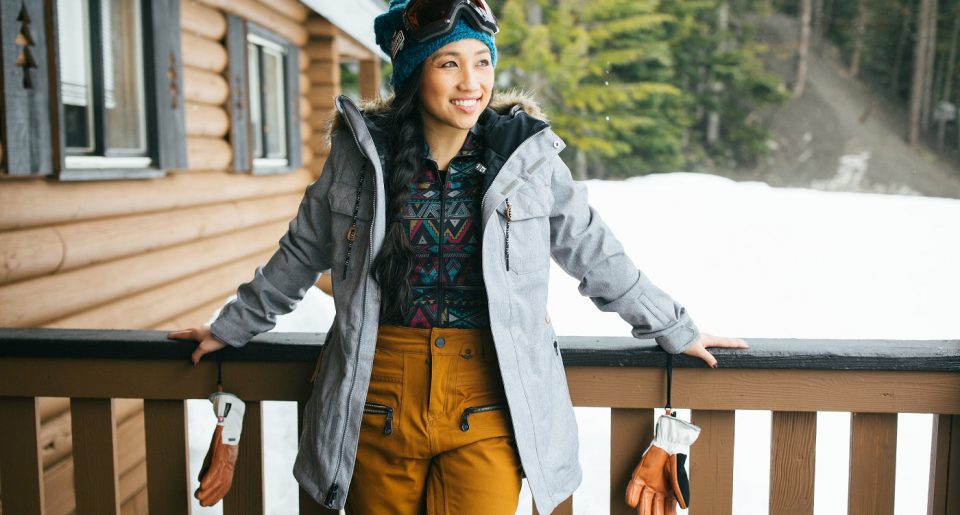 A young female skier wearing colorful gear standing on a covered porch in the snow
