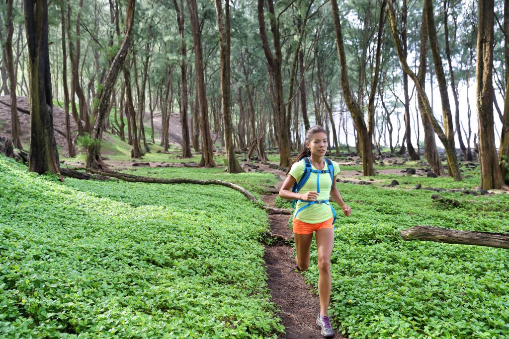 A young female trail runner running through a green forest with an ultra running backpack